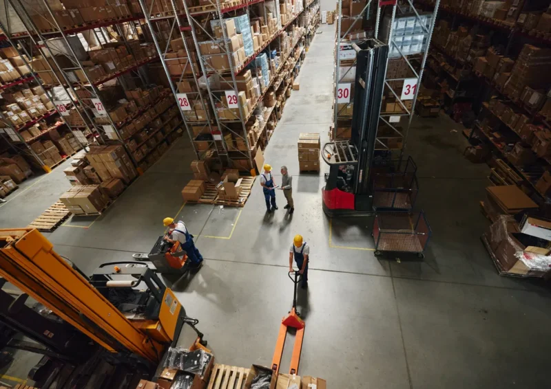 Groot magazijn met medewerkers en heftrucks die werken tussen de stellingen.